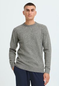 Suéter de punto gris con cuello redondo, mangas largas, puños y dobladillo acanalados, textura suave y un corte holgado, combinado con pantalones oscuros.