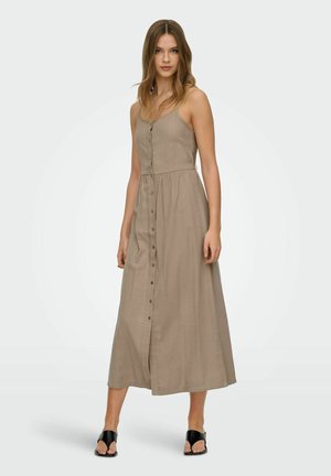Młoda kobieta ubrana w bezrękawnikową sukienkę maxi taupe zapinaną na guziki z przodu oraz czarne sandały z odkrytymi palcami, stojąca na jednolitym tle.