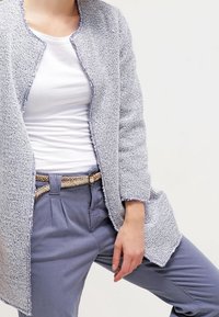 Ljust grått strukturerat stickat cardigan med öppen front, i kombination med en vit tanktop och ljusblå byxor, som har ett flätat bälte.