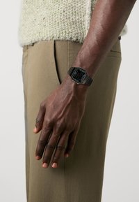 Montre numérique avec un cadran rectangulaire noir et des accents argentés, associée à un bracelet en métal noir, portée sur une main contre un pantalon olive clair.