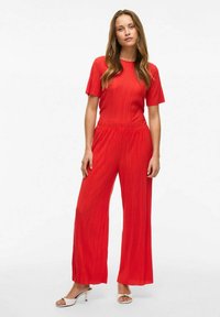 Monos de tirantes rojos con mangas cortas, diseño de pierna ancha, cinturilla elástica y tela texturizada. Combinado con sandalias de tacón blancas.