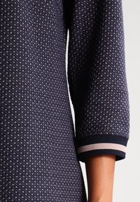 Marinblå stickad tröja med diamantmönster, 3/4 ärmar och marinblå ribbade mudd med en rosa rand.