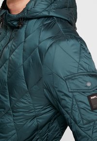 Quiltad teal jacka med huva, med dragkedjestängning, texturerad yta och en ärmklistermärke med en logotyp.