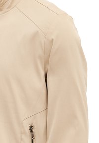 Beige jakke med en glat overflade, strømlinet design, højt krave og en lynlåslomme på siden med metalhardware.
