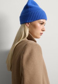 Gorro de punto azul con banda acanalada, con una parte superior puntiaguda. Lleva puesto un abrigo de lana marrón claro, con superficie texturizada visible.