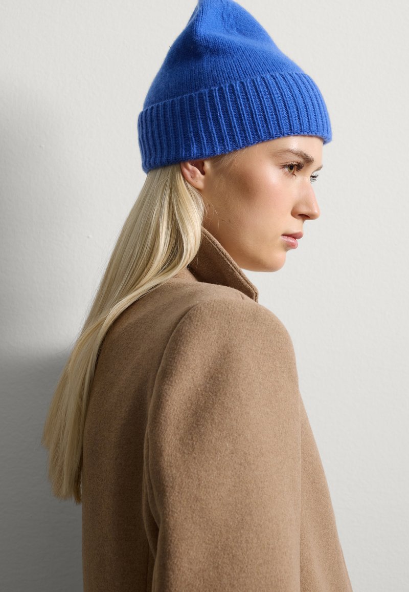 Gorro de punto azul con banda acanalada, con una parte superior puntiaguda. Lleva puesto un abrigo de lana marrón claro, con superficie texturizada visible.