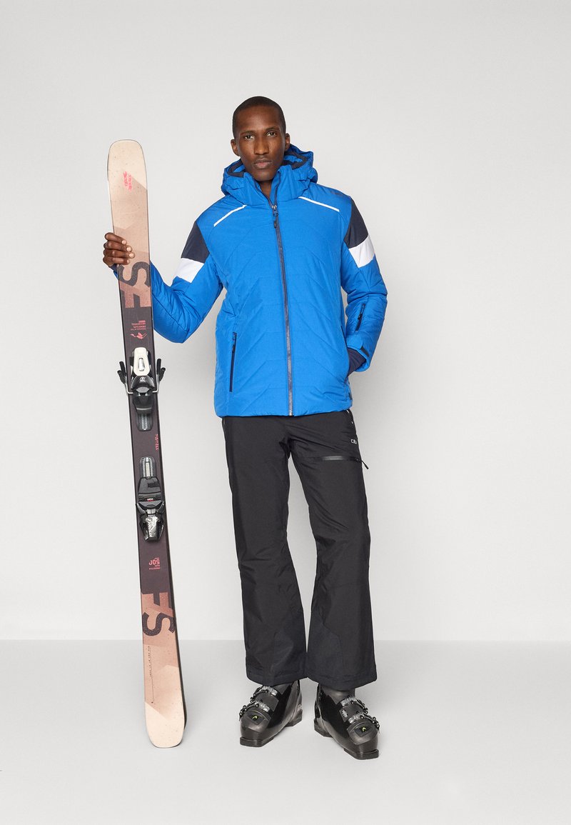 Blauw geïsoleerd skijack met capuchon, zwarte skibroek en skischoenen, terwijl hij een bruine ski met bindingen vasthoudt. Heeft een kleurgeblokte design.