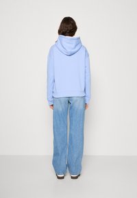 Person som står vänd mot en vägg, iklädd en ljusblå hoodie, vida blå jeans och vita skor på ett enfärgat golv.