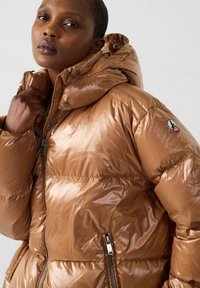 Chaqueta acolchada marrón brillante con capucha. Características incluyen cierre de cremallera, bolsillos laterales y parche con logo en la manga. Diseño acolchado.