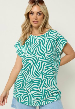 Blond vrouw met een groene en witte abstracte zebrastrip top, lichtblauwe korte broek en een zonnebril op haar hoofd.