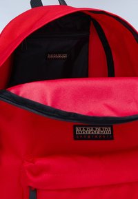 Zaino rosso con una parte esterna in canvas, foderato internamente di nero e dotato di tasca con zip. Presenta un'etichetta con logo e un design strutturato.
