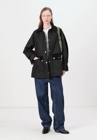 Black quilted jacket with a pointed collar, snap buttons, two front pockets, and gold logo detail. Paired with blue wide-leg jeans and black shoes.