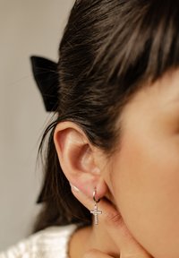 Boucle d'oreille en argent avec une petite croix texturée ornée de strass transparents, portée à l'oreille. L'arrière-plan comprend des cheveux bruns et un ruban noir.