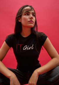 Młoda kobieta z długimi ciemnymi włosami w czarnej koszulce "IT Girl" siedząca na krzyż z nogami na tle czerwonym, patrząca w górę z zamyśleniem.