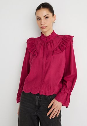 Chemise boutonnée cramoisie avec col et manches à volants, ornée de détails brodés le long de l'ourlet. Associée à un pantalon en jean foncé.