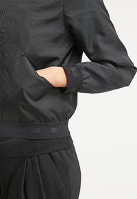 Czarna kurtka bomberka wykonana z gładkiego materiału, z zamkiem z przodu, elastycznymi mankietami, kieszeniami i ściągaczem w pasie.