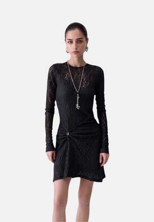 Donna che indossa un abito nero in pizzo a maniche lunghe con dettaglio arricciato e una lunga collana d'argento con un ciondolo a forma di cuore.