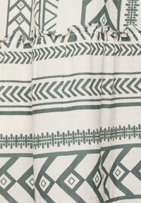 Tkanina bawełniana z kremowym tłem i zielonymi wzorami geometrycznymi, w tym chevronami, diamentami i dekoracyjnymi liniami. Falbany na brzegach.