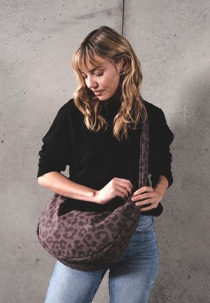 Femeie cu păr blond, purtând un top negru și blugi albaștri, ajustând o geantă gri cu imprimeu leopard în fața unui perete de beton gri.