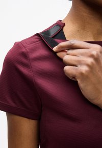 Άτομο που ρυθμίζει μπορντό τοπ με κοντά μανίκια, στερεώνοντας έναν μαύρο ιμάντα βέλκρο κοντά στον ώμο πάνω από μια μαύρη εσωτερική μπλούζα.