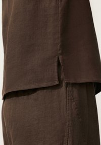 Set din cămașă și pantaloni maro din in, prezentând detaliu de despicătură laterală și cusături la buzunar pe un material ușor.