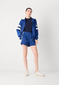 Jaqueta atlética azul com detalhes listrados em branco, combinada com calções azuis e ténis brancos. Tecido liso e leve com fecho de correr.