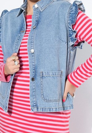 Kvinde iført en langærmet top med pink og røde striber og en lyseblå ærmeløs denimvest med flæseærmegab og forlommer.