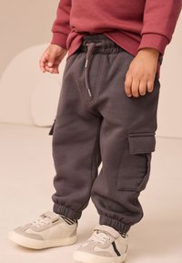 Pilkos laisvalaikio kelnės su elastine juosmens juosta, šoninėmis kargo kišenėmis ir elastiniais rankogaliais, derinamos su pilkais ir baltais sportbačiais.