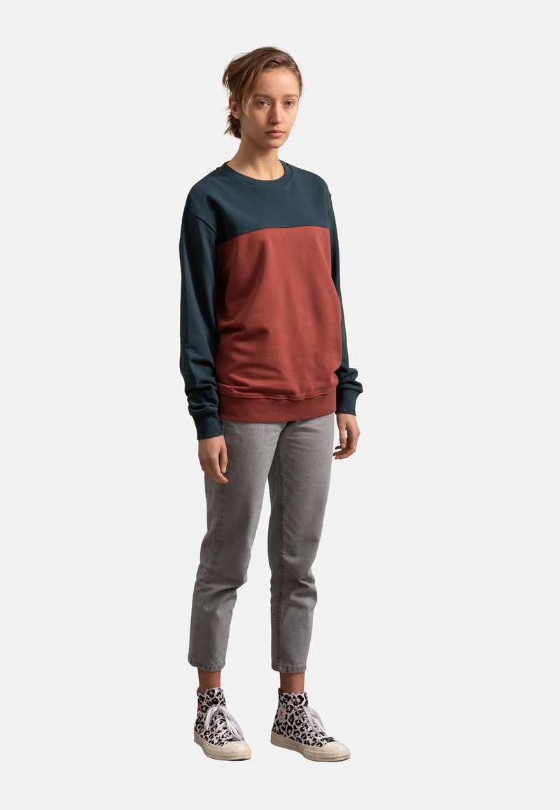 Kleurblok sweatshirt in donker teal en roestkleur, gemaakt van een katoenmix, met een ronde hals en lange mouwen, gecombineerd met grijze broek en gemusterde sneakers.