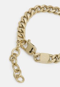 Bracciale in metallo dorato con catena a maglie intrecciate, finitura testurizzata e un robusto gancio a sgancio. Presenta un piccolo tag con dettagli del logo.