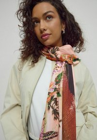 Kohveriku curly tume juustega naine, kes kannab kreemivärvi jakki ja lillemustriga salliga, kus on roosa, oranž ja roheline värvitoon.