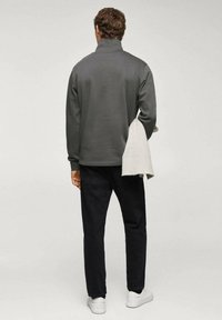 Veste polaire grise à fermeture éclair, pantalon noir, baskets blanches. Le modèle porte un vêtement tricoté gris clair sur l'épaule. Tissu texturé.