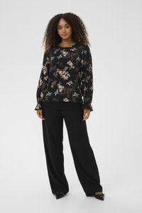 Ženska s kodrasto lasmi, oblečena v črno cvetlično bluzo z dolgimi rokavi, črne široke hlače in črne pete, stoji pred enostavno ozadje.