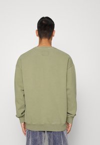 Olivově zelená mikina s dlouhými rukávy, kulatým výstřihem a žebrovanými manžetami. Má čtvercovou visačku na zadní straně poblíž výstřihu.