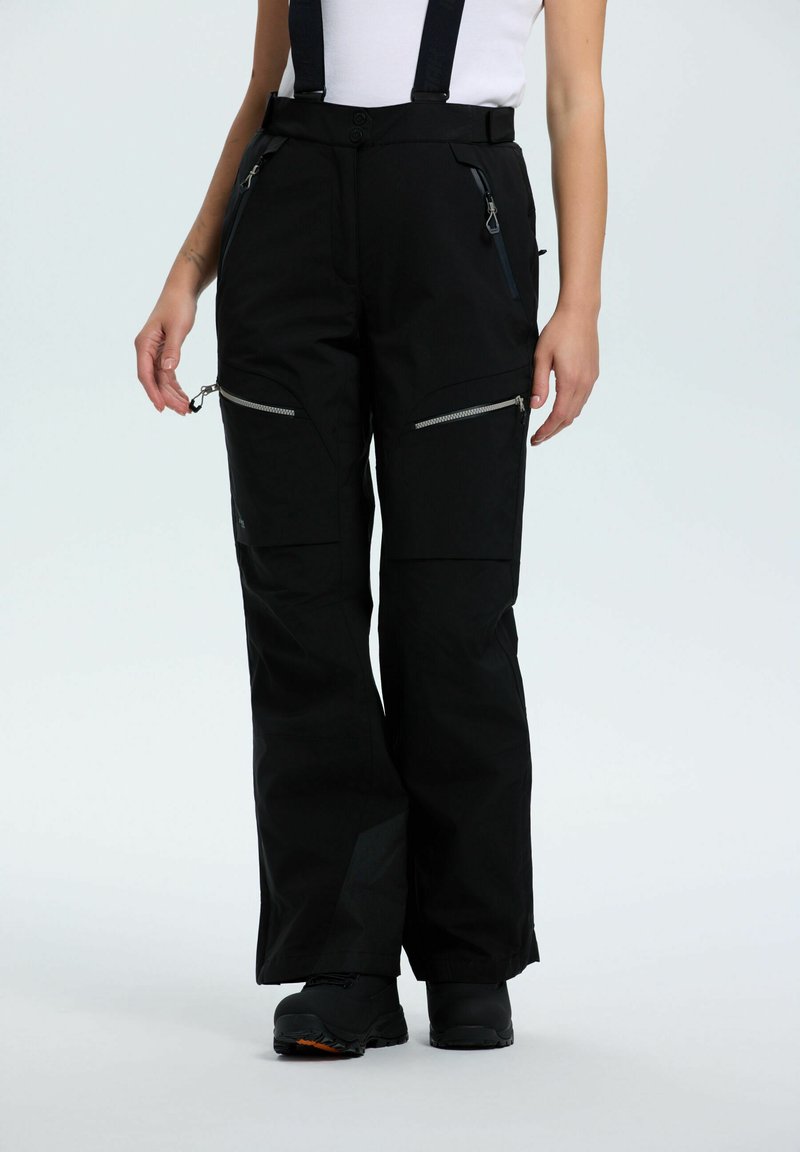 Schwarze Schneehosen mit Hosenträgern, mit Reißverschlüssen an Oberschenkel- und Hüfttaschen, getragen mit schwarzen Stiefeln und einem weißen Oberteil, stehend vor einem schlichten hellen Hintergrund.