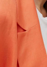 Oranžový lněný blazer s texturovaným povrchem, který má boční kapsu a čistý, pokrývací design. Blazer je oblečený přes bílou košili.