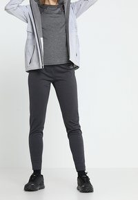 Osoba ubrana w szaro-zamykany sweatshirt na ciemnoszarym t-shircie i czarnych dresach, stojąca z uniesionymi ramionami na białym tle.