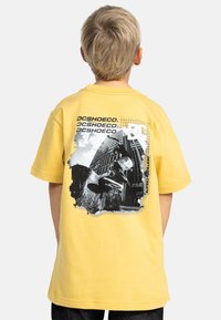 Camiseta de algodón amarilla con un estampado gráfico de un patinador sobre un fondo urbano, logo "DC" y acentos de texto. Corte relajado y mangas cortas.