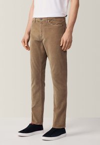 Pantaloni in corduroy marrone con vestibilità slim, dotati di tasche laterali e chiusura con bottone frontale, abbinati a sneakers nere.