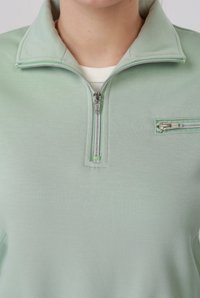 Κοντινό πλάνο ενός ανοιχτοπράσινου πουκαμίσου με φερμουάρ και γιακά, με επιπλέον μικρή τσέπη με φερμουάρ στο στήθος, φορεμένο πάνω από λευκό εσώρουχο.