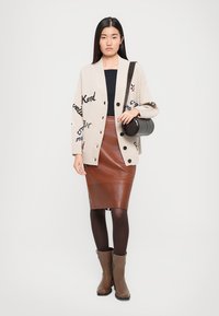 Cardigan di maglia beige con dettagli testuali, top nero, gonna a matita in pelle marrone, collant scuri e stivaletti alla caviglia marroni, mentre tiene in mano una borsa rotonda nera.