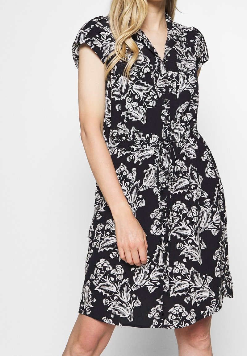 Robe noire à motif floral blanc, manches courtes, boutonnée sur le devant, ceinture à nouer à la taille, longueur genou, en tissu léger.