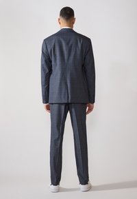 Giacca e pantaloni a scacchi grigio scuro, design aderente, tessuto texture, abbinati a sneakers bianche. Visti da dietro.