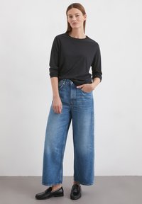 Juodas ilgas rankovėmis marškinėliai derinami su aukštu liemeniu, plačiais mėlynais džinsais ir juodais loafers batais. Lygus audinys ir laisvas siluetas. Minimalistinis dizainas.