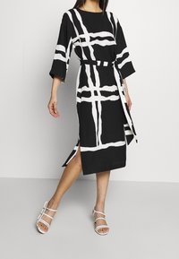 Abito nero con motivo a griglia bianca, maniche larghe, vita leggermente cintrata con cintura, fessure laterali e abbinato a sandali bianchi con cinturini. Tessuto dalla texture liscia.