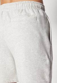 Pantalones cortos deportivos de color gris claro, fabricados en un tejido suave, con una cintura elástica y un bolsillo lateral. El texto en la pierna incluye los detalles de la marca y un lema.