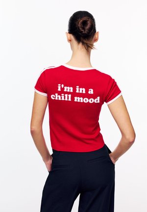 Femme aux cheveux attachés en chignon, portant un t-shirt rouge avec le texte blanc "je suis d'humeur détendue", debout les mains dans les poches d'un pantalon noir.