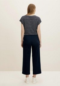 Kvinna med ljust brunt hår som bär en kortärmad randig topp, mörka vida byxor och vita högskor, stående mot en enfärgad bakgrund.