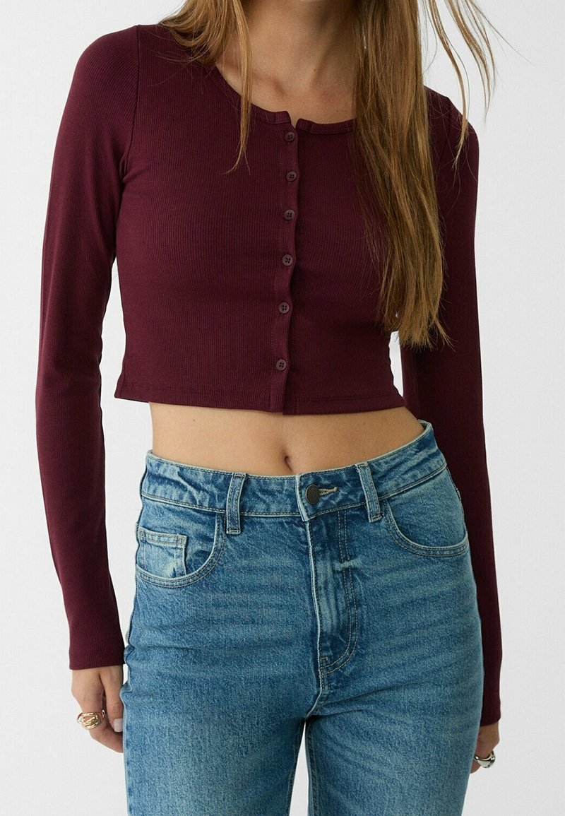 Femme portant un top court bordeaux à manches longues et boutonné, et un jean bleu taille haute, debout devant un fond uni.