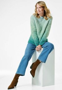 Strickpullover in grünen Farbverläufen, kombiniert mit blauen Jeans und braunen Wildleder-Ankle-Boots. Das Model sitzt auf einem weißen Sockel.